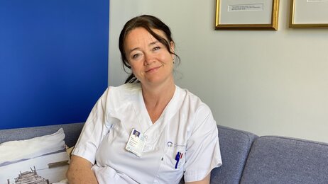 Bildet viser Aina Evensen Helme på jobb i sykepleieruniform. Hun sitter og ser inn i kameraet