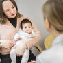 Bildet viser en mor med et spedbarn på fanget. Hun sitter på en stol overfor en helsesykepleier, som vi ser ryggen på