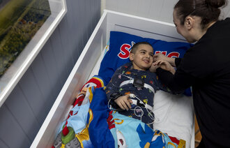 The photo shows a boy lying in his bed. Tools for sleep testing and monitoring are equipped on the boy. His mother is bending towards him, both are smiling