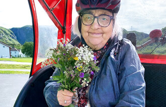 Bildet viser en eldre som sitter i en sykkeltilhenger ute i naturen. I hånden holder hun en bukett markblomster.