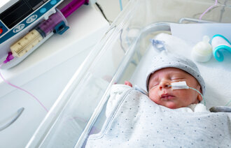 The image shows a baby lying in a hospital bed. The baby has a feeding tube through the nose, and a syringe containing breast milk is placed beside the baby.
