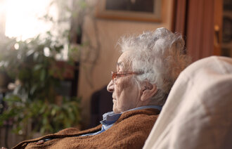 Bildet viser en eldre dame som sitter ved et vindu og kikker ut