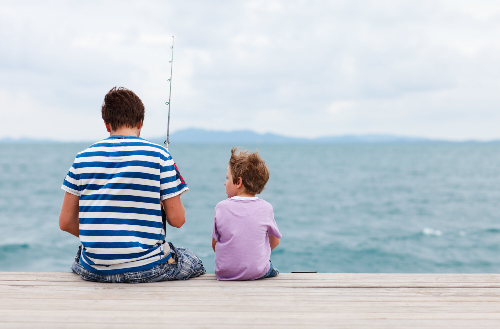Et lite barn sitter sammen med en voksen mann på en brygge. Mannen fisker. Et lite barn sitter sammen med en voksen mann på en brygge. Mannen fisker.