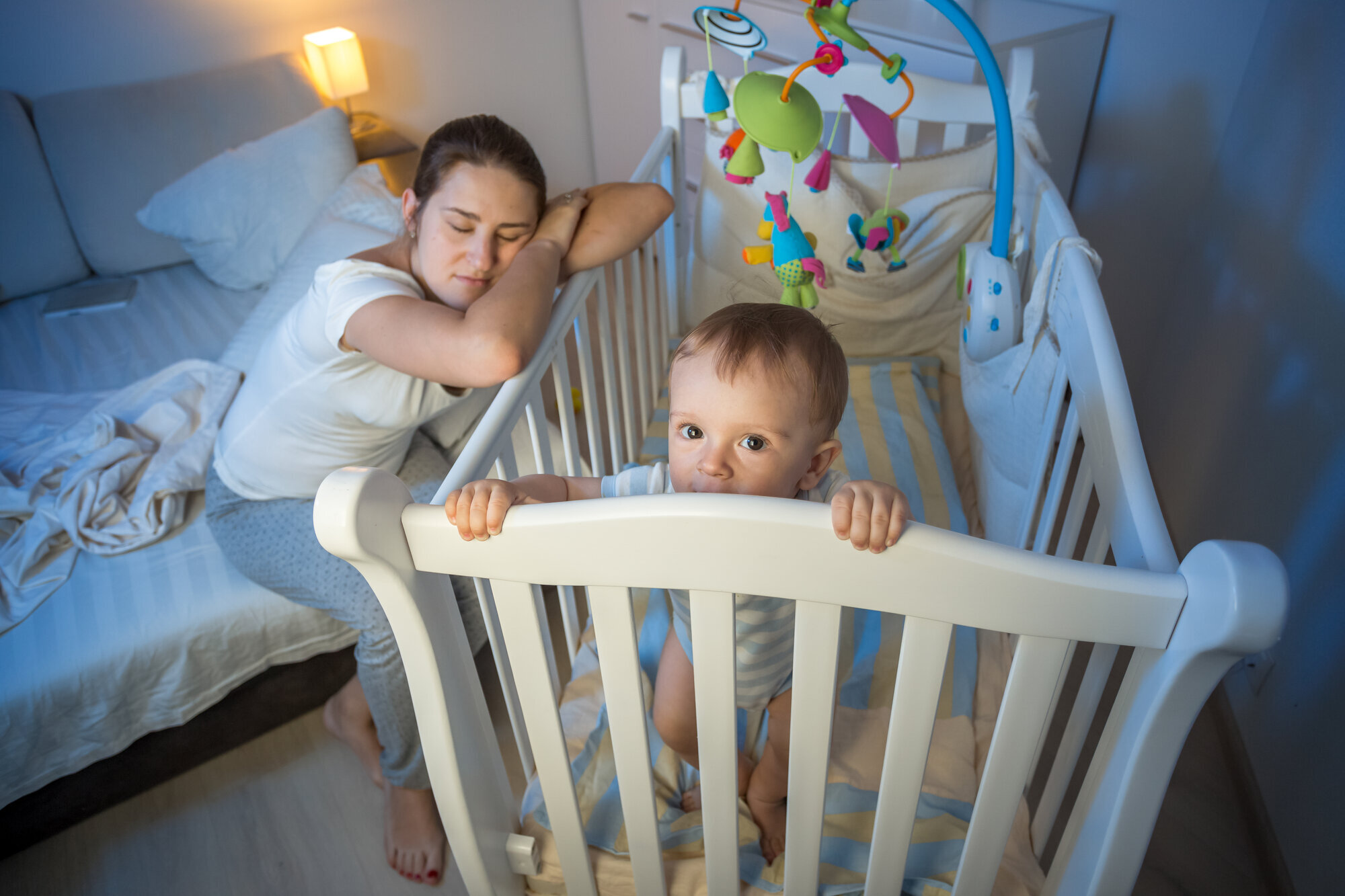 Trøtt mor og våkent barn Bildet viser en mor som halvsover over barnesenga, mens barnet står i senga, lys våkent