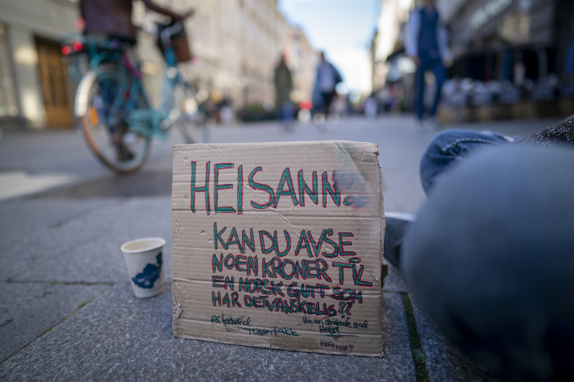 Oslo Bildet viser Karl Johans gate og en plakat som en sittende mann har foran seg, hvor det er bønn om bidrag til en som har det vanskelig.