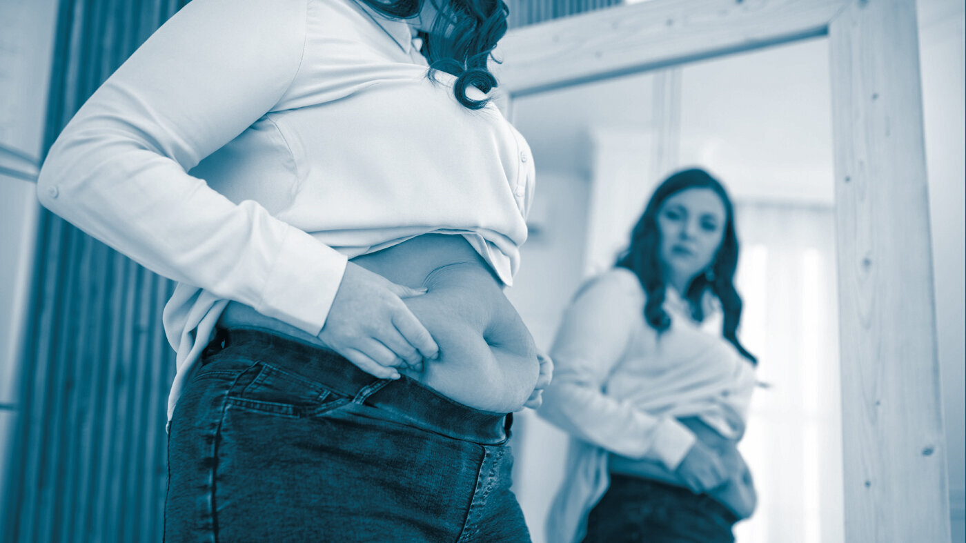 Overweight woman The photo shows a woman looking in the mirror. We can see her belly, and she is squeezing it to show she has a couple of kilos extra