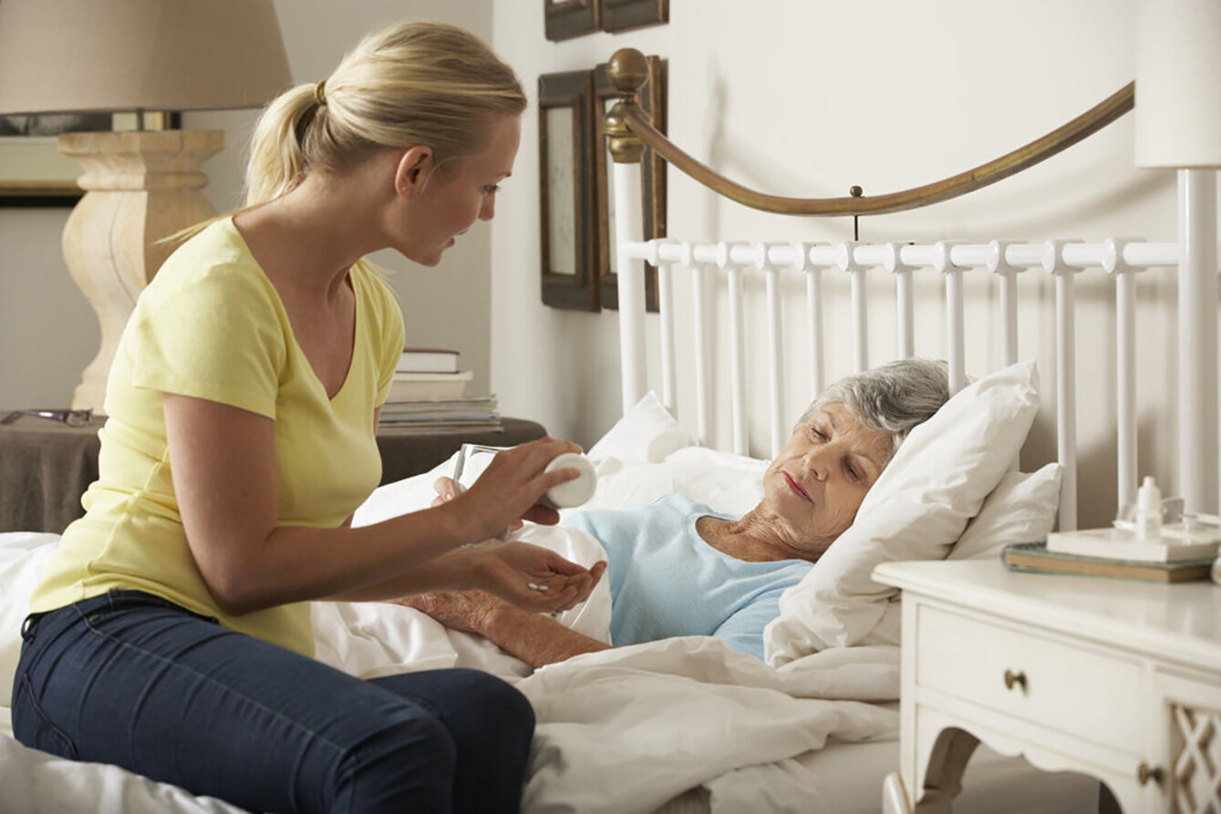 Bildet viser en eldre, syk dame i senga. På sengekanten sitter dattera