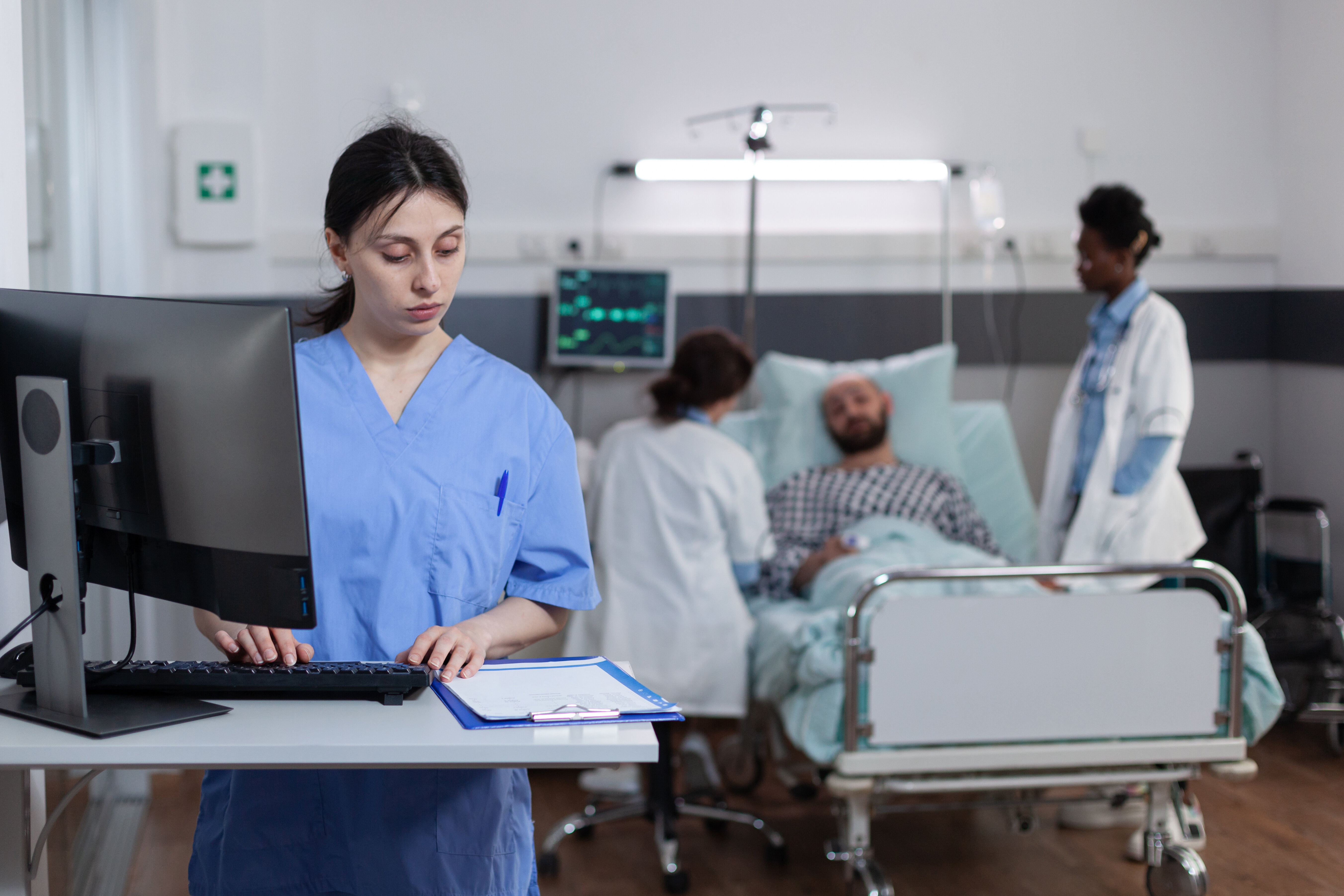 The image shows a nurse standing at a computer in a patient room. A form is placed in front of her. In the background, a patient is lying in bed while two nurses stand at the bedside.