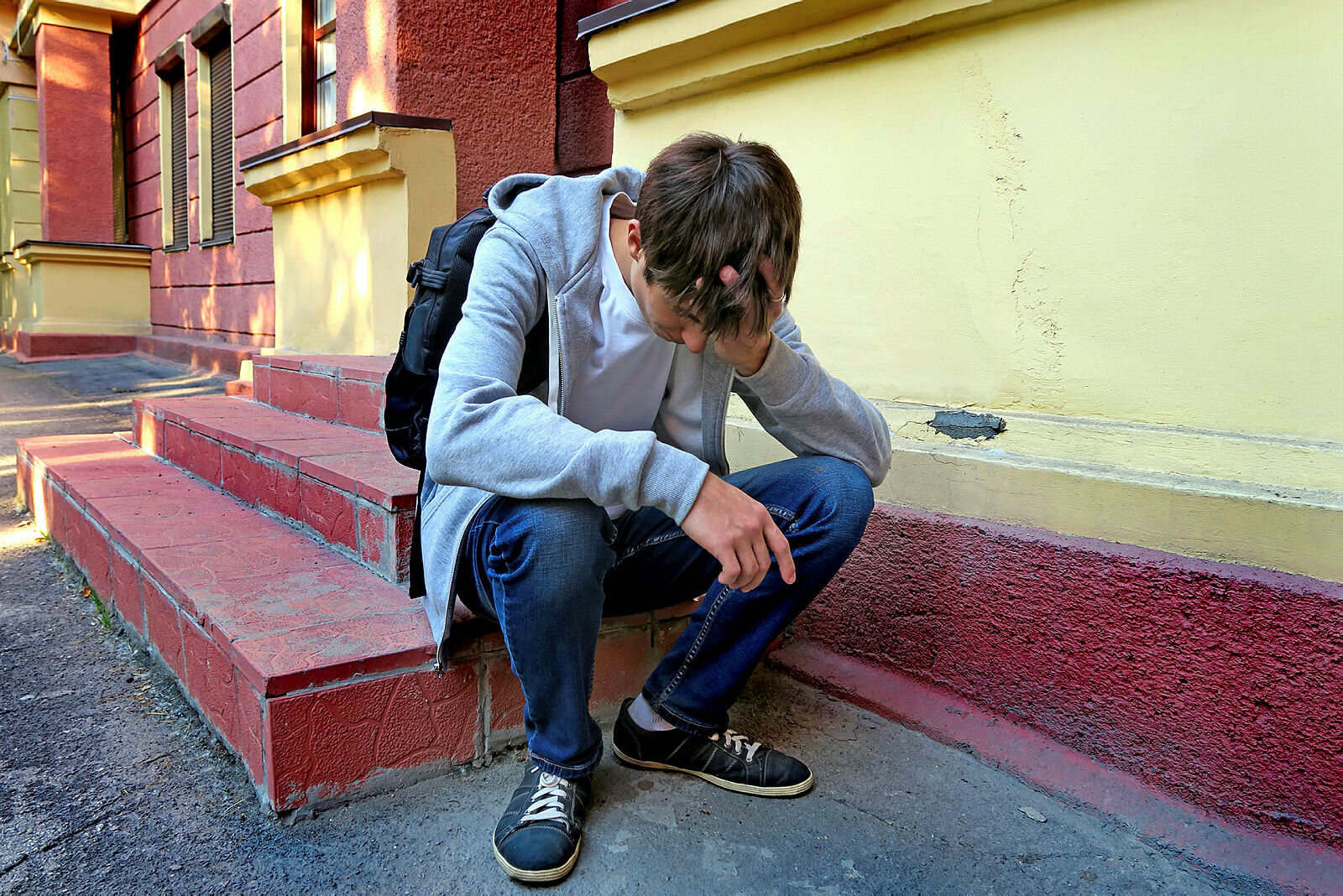 Tenåring sitter på en trapp utenfor skolen Bildet viser en tenåringsgutt med ransel som sitter på en trapp utenfor skolen. Han er lei seg.