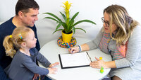 The photo shows a father with his daughter on his lap.  They are sitting at a table opposite a woman leaning towards them. She has a notebook in front of her.