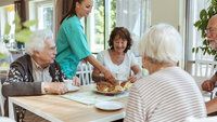 Beboere og sykepleier på sykehjem Bildet viser fire eldre som sitter rundt et bord. En sykepleier serverer rundstykker til dem