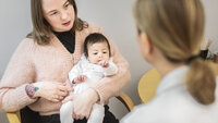 Bildet viser en mor med et spedbarn på fanget. Hun sitter på en stol overfor en helsesykepleier, som vi ser ryggen på