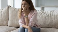 Ettertenksom kvinne Bildet viser en ettertenksom, ung kvinne som sitter i en sofa. Hun ser litt fortvilet ut og støtter hånda halvveis foran munnen