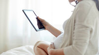 Gravid med nettbrett Bildet viser en gravid kvinne som sitter i en seng og ser på et nettbrett